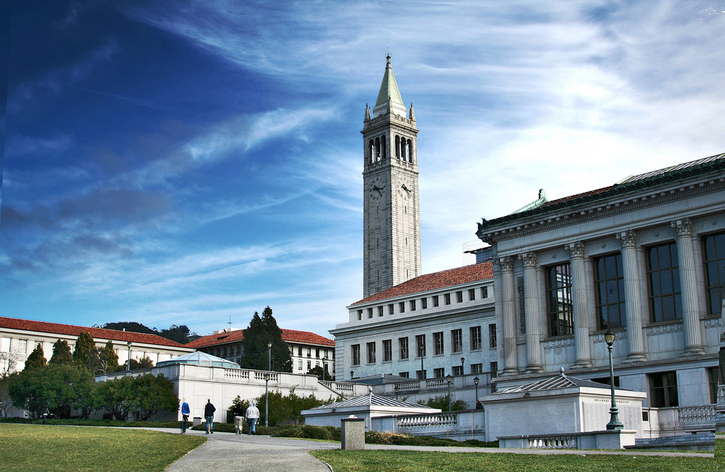 Four people walk through Memorial Glade in front of Doe Library on UC Berkeley's campus. The Campanile towers in the center of the background.