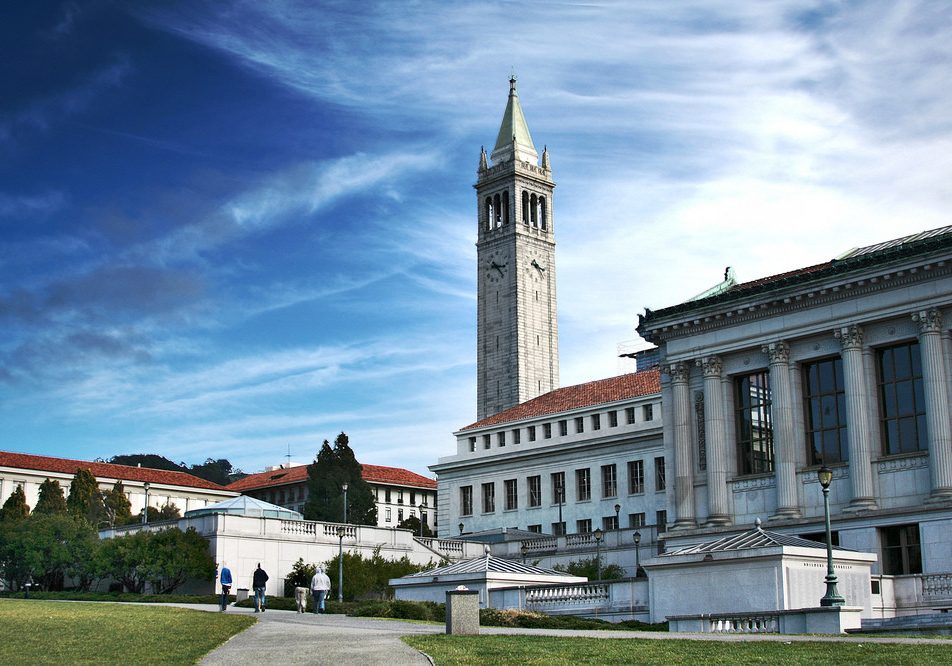 Four people walk through Memorial Glade in front of Doe Library on UC Berkeley's campus. The Campanile towers in the center of the background.