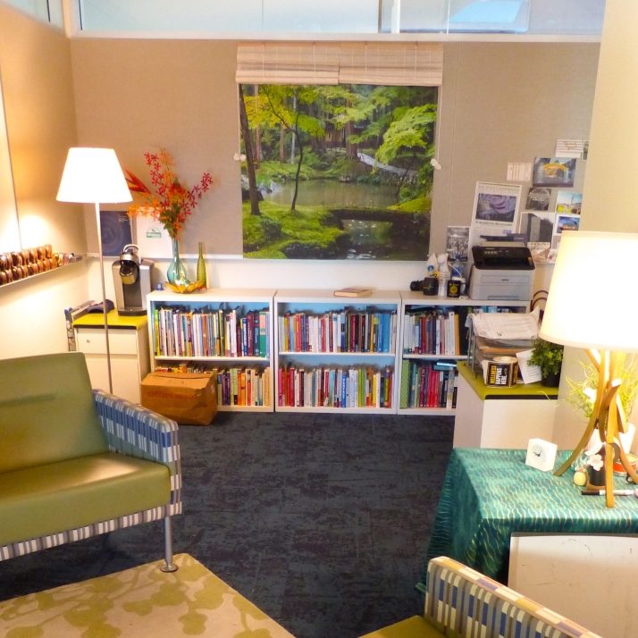A warmly lit room with two green arm chairs, a low bookshelf, and a hanging canvas depicting a green forest.